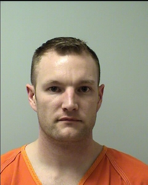 A man with short light brown hair wearing an orange prison jumpsuit is shown facing forward against a plain light-colored background.