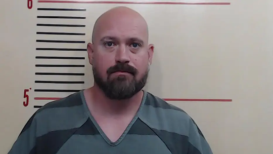 A smiling woman stands in front of a black door, holding flyers and a blue drawstring bag—part of the Field Team in West Virginia. She wears a teal jacket, blue pants, and sneakers. Green shrubs and a brick porch are visible.