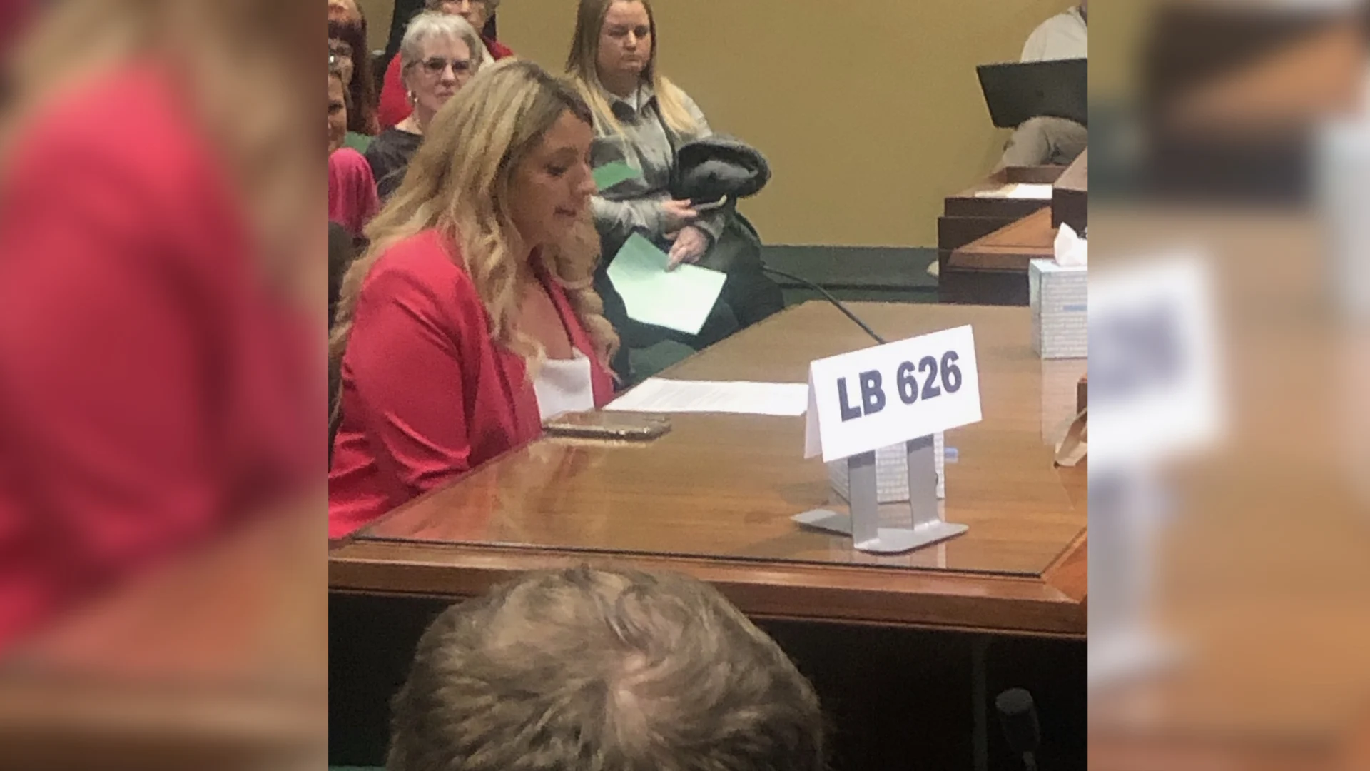 A woman in a pink blazer, identified as Haile, speaks at a hearing about abortion drugs, seated at a wooden table with a sign labeled LB 626. Several people sit and listen in the background.